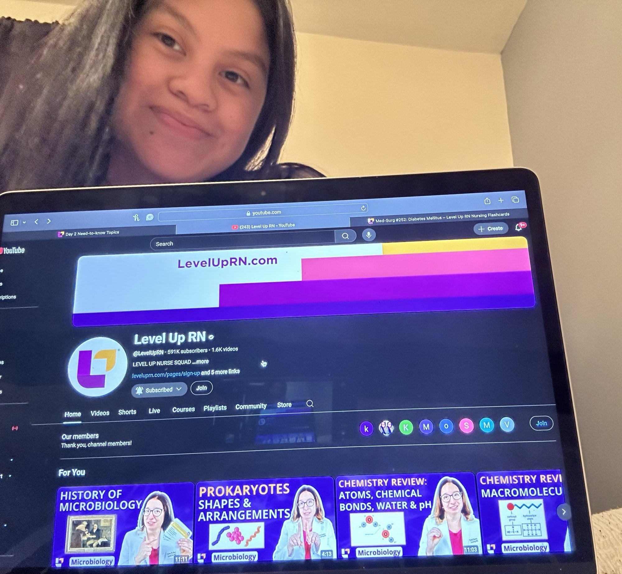 Emily holding up a laptop showing the Level Up RN Youtube homepage with new microbiology videos
